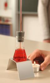 The image shows a test tube filled with red liquid resting on a sturdy stand. Next to the tube is a burning tea candle while a hand holds the tube, depicting an educational experiment setup.