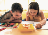 The picture shows two children sitting at a table, eagerly observing an experiment with a glass carafe containing water and colorful figures. Around them are other experiment materials and notepads, which suggests a learning story in