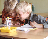 The picture shows two children who are curiously conducting an experiment. In front of them is a device with a yellow ball, while they are concentrating on a sheet of paper with instructions.