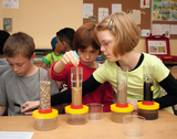 The picture shows children experimenting with different containers in a classroom. They appear to be conducting an experiment in which they layer different materials into transparent cylinders and observe them.