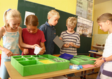 The picture shows several children standing at an experiment table, working with various materials from a green foam box. They appear to be focused as they work on their projects in the classroom.