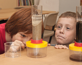 The picture shows two children looking fascinated at an experiment kit that appears to show different layers of materials. The kit consists of transparent containers containing differently colored substances and
