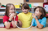 The picture shows three children holding two glass flasks filled with water, connected by a tube. They are experimenting and probably observing a physical phenomenon, while a friendly learning atmosphere
