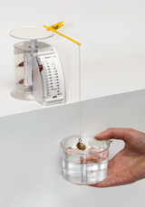 The picture shows a weighted levitation device used to determine the buoyancy of a body in water. A cup of water is held in the hand while a measuring scale and a spring balance are used for accurate weight analysis.