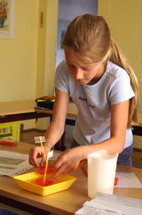 The picture shows a girl working concentratedly on an experiment using a yellow bowl with red liquid and several labelled sticks. In the background you can see tables and a classroom atmosphere that is typical for school