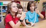 In the picture, three children are experimenting with water in glass containers and a hose. They seem to be watching with interest how the water in the containers acts, which promotes interactive learning.