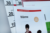 The picture shows a teaching material entitled &quot;Heat&quot; from Cornelsen Experimenta, which focuses on experiments on the topic of heat. In the background, a thermometer is visible, showing different degrees of temperature.