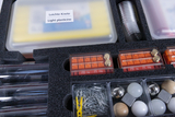 The picture shows a sorted experiment box with various materials, including modeling clay, measuring cylinders, weights and various components such as balls and pieces of wood. The contents are clearly arranged in compartments, ideal for school experiments.