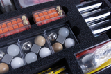 The picture shows a sorted experiment box with various materials, including balls made of wood, metal and plastic as well as measuring cylinders. The compact arrangement of the contents is housed in a foam insert that has an orderly groove