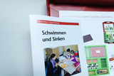 The picture shows a brochure entitled &quot;Floating and Sinking&quot; from an experiment set for the educational sector. It shows children experimenting and learning together with a teacher at a table.