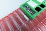 The picture shows an arrangement of test tubes in a red holder and a green set of laboratory equipment, including pipettes and plates. In the foreground is a container with a label for chemicals.