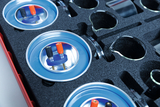 The image shows a textured foam tray containing various experimental devices and accessories. The focus is on round holders with colored terminals and other glass containers used for educational experiments.