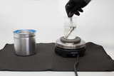 The image shows a laboratory setup with a beaker containing water placed on a heater, while a gloved arm inserts an element into the water. Next to the heater is a metal container, probably for experimentation.