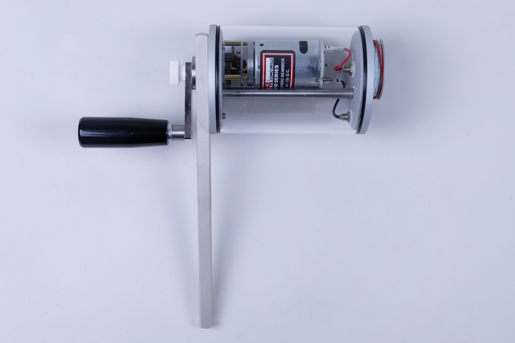 The image shows a transparent device with a handle and a motor inside. It appears to be designed for educational experiments, possibly to demonstrate mechanical motion or energy conversion.