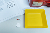 The picture shows a yellow experiment container, a small magnet and a round, white disk, which probably come from an experiment kit. In the background is a textbook with illustrations and explanations of magnets and compasses.
