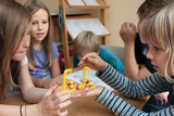 The picture shows several children concentrating on experiments at a table. They are holding two yellow experiment boxes with a pendulum and appear to be conducting a physical investigation.