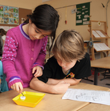 The picture shows two children working on an experiment. A girl wearing a purple sweater is dipping a small object into a yellow bowl while a boy looks on with interest and instructions lie on the table.