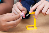 The picture shows two children conducting an experiment together. They use a yellow support element and a paper clip to manipulate a piece of checkered material.