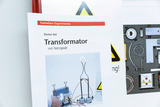 The picture shows instructions for a demo set on the topic of transformer with mains coil. In the background you can see various experimental devices and safety warnings that indicate experimental use.
