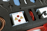The picture shows a box with scientific experiment equipment, including two devices with electrical connections and warning symbols. In addition, various cables and other instruments are housed in foam moldings for safe storage.