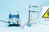 The picture shows an experimental device with two high voltage power sources used for educational purposes. Next to the devices there is a warning sign indicating the high voltage.