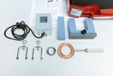 The image shows an arrangement of experimental materials, including a transformer, various holding devices and a heating coil, typically used in educational settings. In the background, technical documents relevant to conducting experiments can be seen.