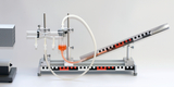 The picture shows an experimental device with a slanted tube that leads a liquid into a transparent chamber. Next to the tube are various connections and an electrical device that presumably serves to control the experiment.