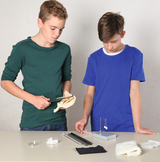 The picture shows two young people working at a table with various experimental materials. One of the boys is holding an instrument, while the other is focused on a physics experiment with a spring balance.