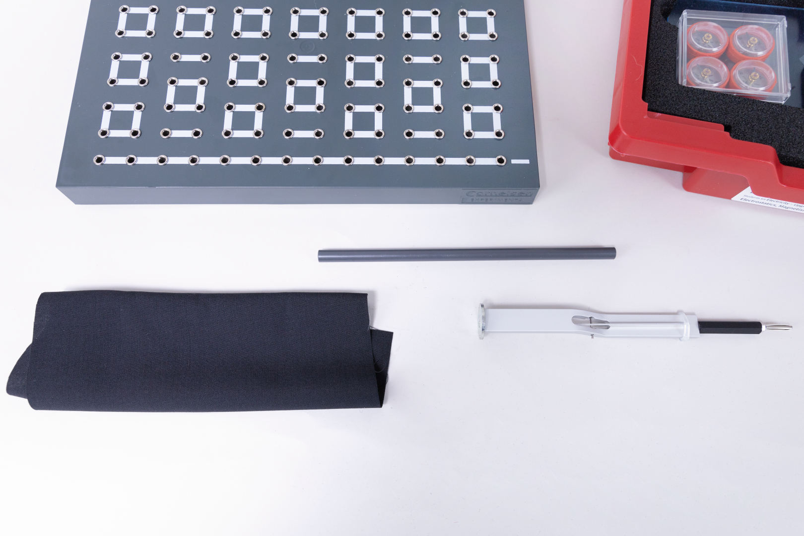 The picture shows a selection of experiment materials, including a plate with holes, a black cloth, a grey stick and a pen. These materials are intended for use in education and support creative and scientific experiments.