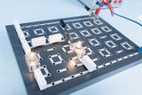 The picture shows an experimental board with various connection options, on which two light bulbs are lit. Two cables are connected that create an electrical connection, and the arrangement can be used for various experiments in the educational field.