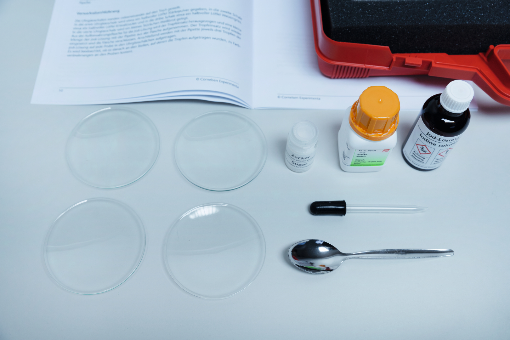 The picture shows various laboratory equipment, including two petri dishes, chemicals such as sugar and iodine solution, as well as a dropper and a spoon. In the background is an information booklet, presumably containing instructions for experimental setups.