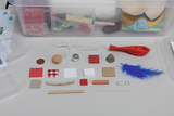 The picture shows a collection of different materials that can be used for experiments. These include pieces of fabric, wood, a marble, a balloon, and other objects in different colors and shapes that serve educational purposes.