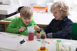 The picture shows two children working together in an experiment room. They examine a container of red liquid and a jar of soil and plants while interacting with excitement and curiosity.