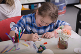 The picture shows a child working concentratedly at a table and drawing on a white surface with various pens. Next to the child, there is a glass with flowers and other craft supplies on the table, indicating a creative activity.