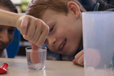 The picture shows a child looking fascinated at a sleight of hand experiment in which a finger is dipped into a glass of water. In the background, other children can be seen watching with interest.