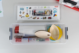 The picture shows a transparent storage box with various experimental materials, including wooden spoons, colorful plates and sample containers. On the top of the box there is a lid with pictures of the materials contained for orientation.