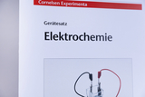 The picture shows a product from an online shop for experimental supplies, especially for the topic of electrochemistry. It illustrates a set of equipment with a picture of an electrolysis experiment, including a transparent cell and cables in black and red.