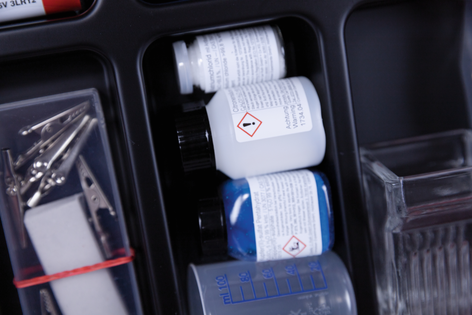 The picture shows a selection of chemicals in bottles arranged in an experiment kit. There are also measuring instruments and other laboratory equipment that can be used for school experiments.