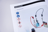 The image shows a page from an electrochemistry textbook that deals with the topic of electroplating. Safety and danger signs are shown, as well as illustrations of experimental materials such as an electrochemical cell and accessories.