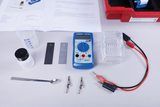 The picture shows various experimental materials, including a multimeter, a beaker, metallic plates, soldering tongs and a spoon. These utensils are typical for chemical or physical experiments in educational settings.