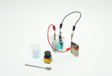 The picture shows an experiment set with an electrolysis device, consisting of a transparent chamber with blue liquid, two cables, a battery, a measuring cup and a test tube. Next to it is a container with chemical reagent and a spoon.