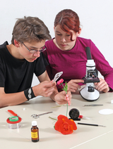 The picture shows two students experimenting at a table. The boy is looking at a flower through a magnifying glass, while the girl is working on a microscope to analyze details of the samples, surrounded by various tools and materials for their experiments.