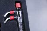 The picture shows a modified glass container with two white outlets and red caps, which are placed in a padded foam insert. This device is probably used for experiments in the educational sector to transport or combine different liquids.