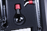 The picture shows a set of laboratory equipment housed in a protective foam insert. It includes a round flask with a red stopper, various glass tubes and a spiral used for chemical experiments.
