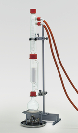 The picture shows a chemical experiment device consisting of several glass containers and tubes mounted on a stable base. There is a heat source at the bottom, while the setup can be used to carry out chemical reactions or distillations.