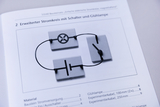 The picture shows a schematic representation of an extended circuit with a switch and a light bulb. The connecting elements are arranged graphically and are used to illustrate electrical basics in education.
