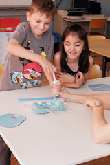 In the picture, children are doing a science experiment in a classroom. A boy is using a syringe while a girl looks on intently, and materials are in a plastic bag on the table.