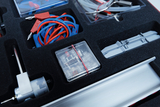 The picture shows an experiment box with various components, including cables in blue and red, a small motor and some transparent components. Everything is neatly arranged in a black padded compartment that ensures safe storage.