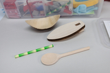 The picture shows various experimental materials, including a semi-circular wooden bowl, an elongated wooden body and a wooden spoon. In addition, a green patterned paper strip lies on the table, surrounded by other craft materials in the background.