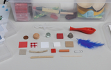 The picture shows a selection of different experimental materials intended for use in education. These include materials of different colors and textures such as wood, fabric, plastic and metals, arranged on a neutral background.